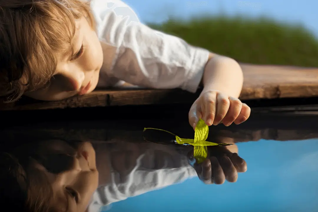 Enfant jouant au bord de l’eau avec une feuille verte.