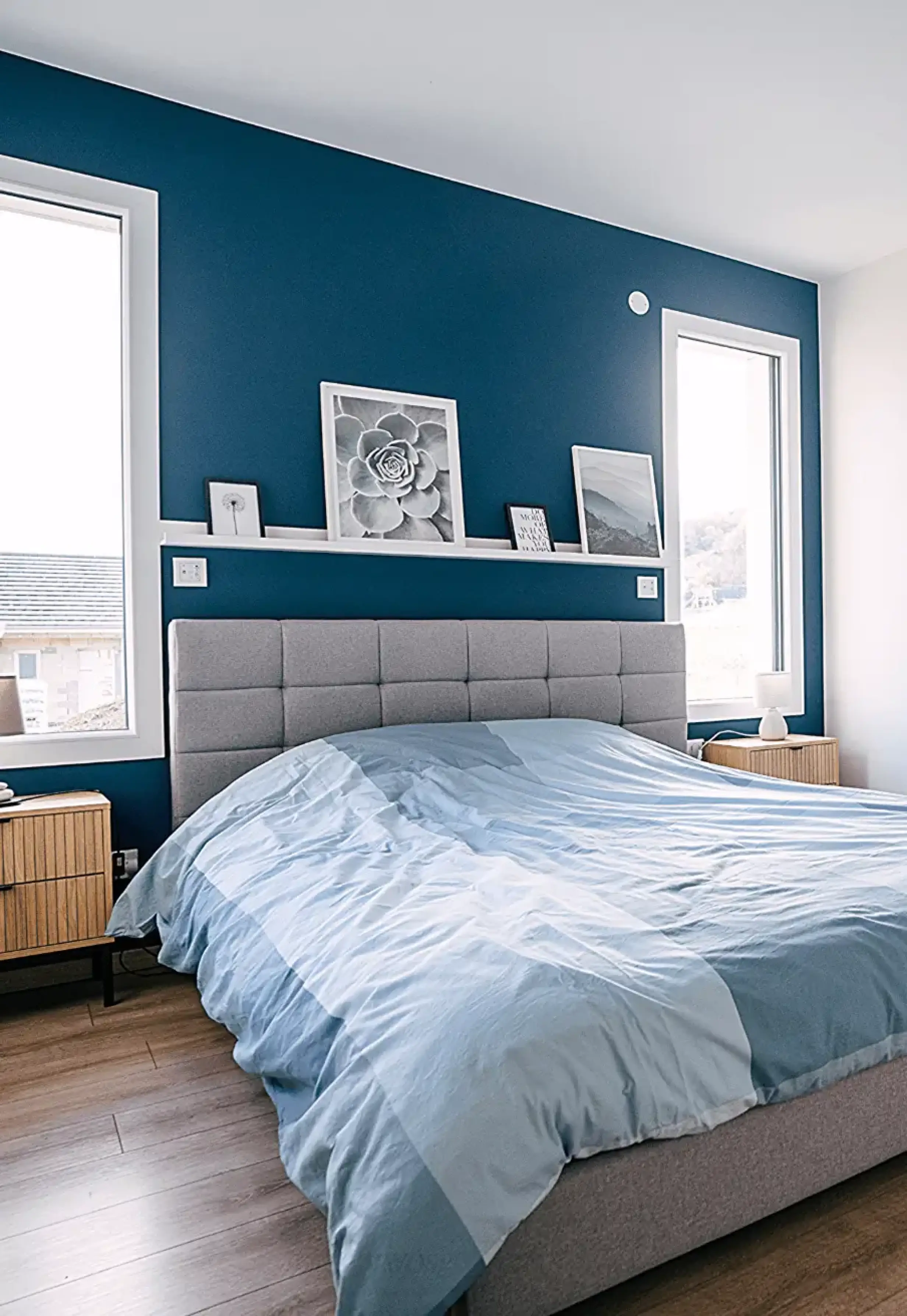 Chambre parentale moderne avec tête de lit grise, murs bleu foncé et salle d’eau attenante.